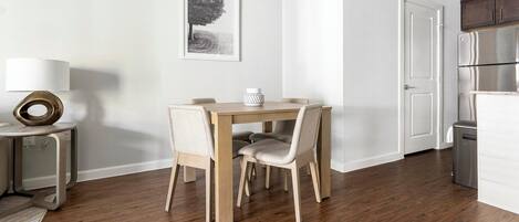 A dining area with a wooden table and chairs, a small side table with a lamp, a framed picture on the wall, and a kitchen area with a refrigerator and cabinets.