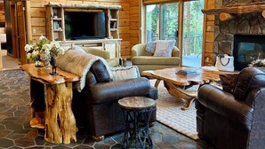 A spacious living area with a stone floor, wooden walls, and a stone fireplace. There are two leather sofas, a coffee table, and a television. - Luxury Mountain Cabin: Hot Tub, Mtn Views, Game Room - Sleeps 10 - Winter Park (Fraser)