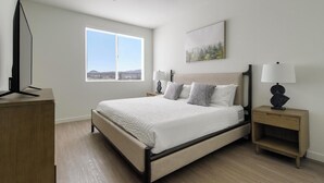 A modern bedroom with a large bed, two bedside tables with lamps, a flat-screen TV on a wooden stand, and a window with a view of the outdoors. - Landing | Chic 2BD, Pool, Gym (Spring Valley)