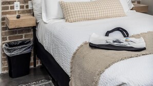 Une chambre avec un mur en briques, un lit avec une literie blanche, un coussin à carreaux, une poubelle noire et une lampe fixée au mur.