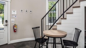 Une pièce avec une table ronde en bois, deux chaises noires, un extincteur et un escalier avec une rampe noire.