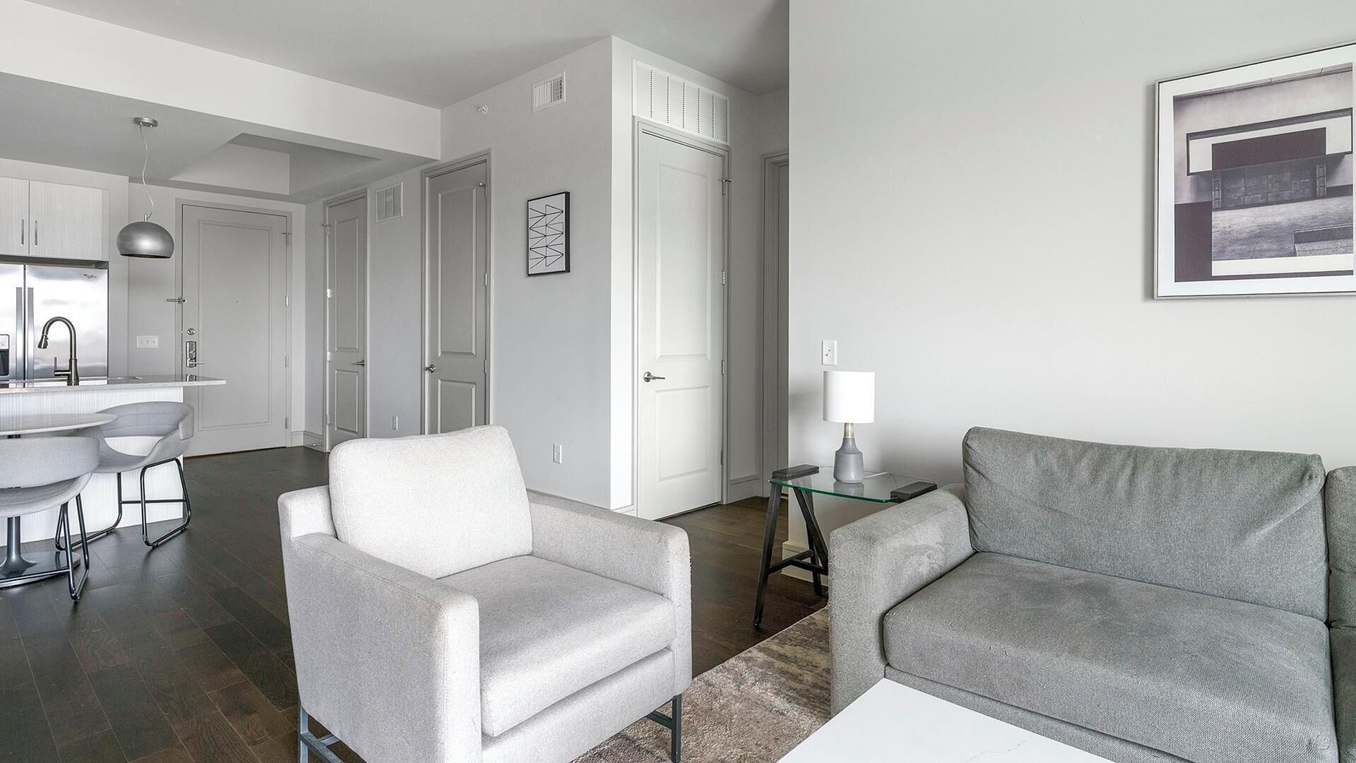 A modern living room with a grey sofa, a white armchair, and a glass coffee table. There is a kitchen area with a white countertop and a sink visible in the background.