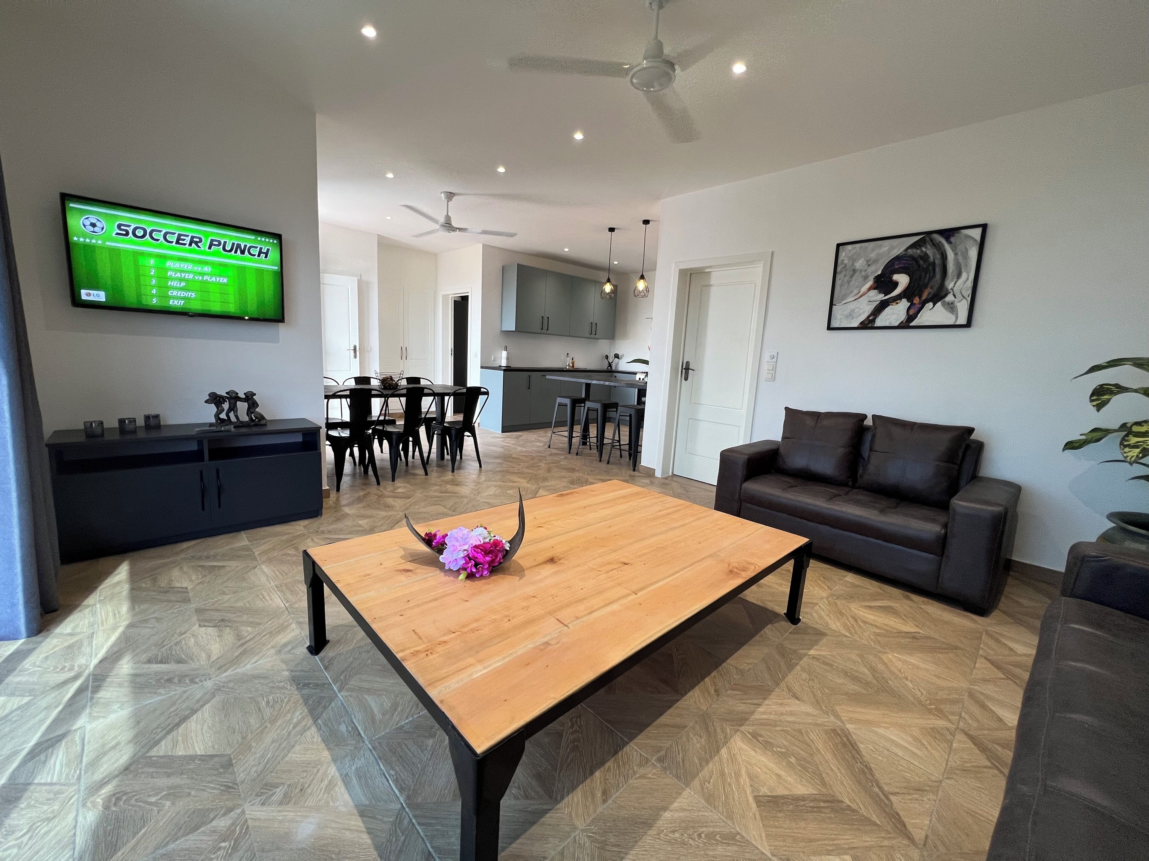 A modern living room with a wooden coffee table, a black sofa, a TV displaying a soccer game, and a dining area with a black table and chairs.