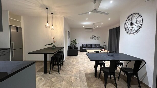 Eine moderne Küche mit Einbaukühlschrank, ein Essbereich mit schwarzem Tisch und Stühlen sowie ein Wohnbereich mit Sofa und dekorativer Wanduhr.