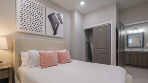 Quarto arrumado com cama, travesseiros, mesa de cabeceira, uma obra de arte na parede, uma porta e banheiro com espelho e pia.