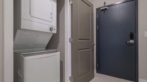 A laundry room with a front-loading washer and dryer, a door with a peephole, and a light switch on the wall. - Landing | Spectacular 1BD in Fountain Heights (Birmingham)