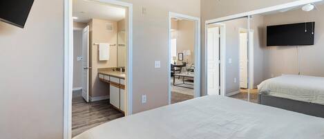 A hotel room with a large bed, a desk with a chair, a television mounted on the wall, and a door leading to another room.