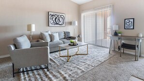 Un salon moderne avec un canapé gris, une table basse et un bureau avec une chaise. On y trouve deux lampes, une grande fenêtre avec des stores et des œuvres d’art accrochées aux murs.