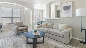 A modern living room with a beige sofa, a round coffee table, and a chair. There are large windows with blinds, a lamp, and a framed picture on the wall. - Landing | Beautiful 1BD, Gym, Pool (Houston)