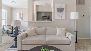 A living room with a beige sofa, a coffee table, and a dining area with a table and chairs. - Landing | Beautiful 1BD, Gym, Pool (Houston)