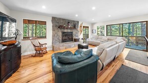 A modern living room with a stone fireplace, wooden flooring, a leather sofa, a white couch, and a large window with a view of trees. - Secluded Incline Village Stay w/ Deck Near Diamond Peak & Lake Cloud 9 and 1/2 by Avantstay (Incline Village)