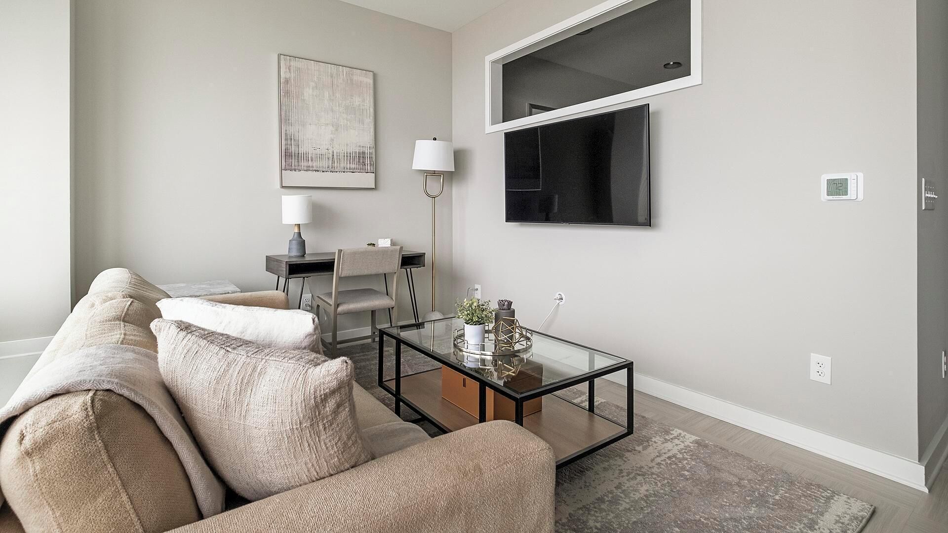 Un salon moderne avec un canapé beige, une table basse en verre, un bureau avec une lampe et un grand téléviseur fixé au mur.