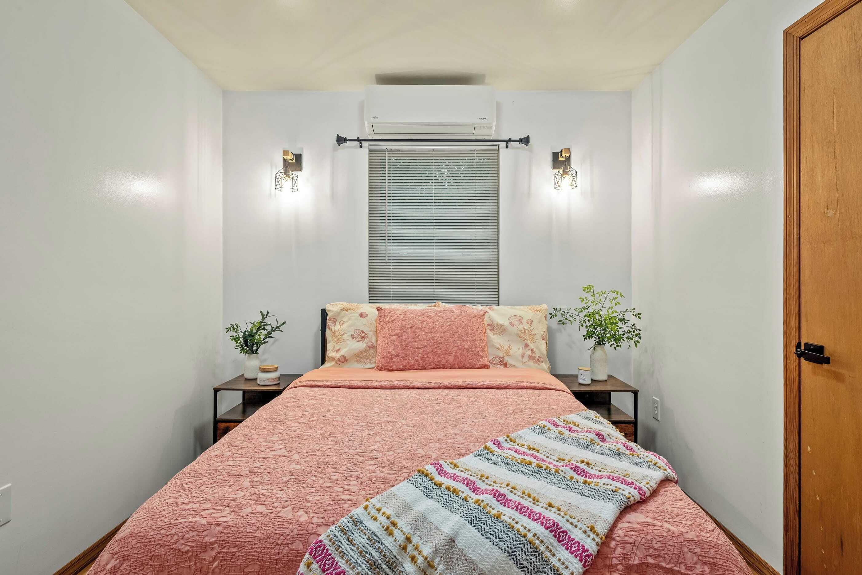A bedroom with a bed, a window with blinds, two bedside tables with lamps, and a door.