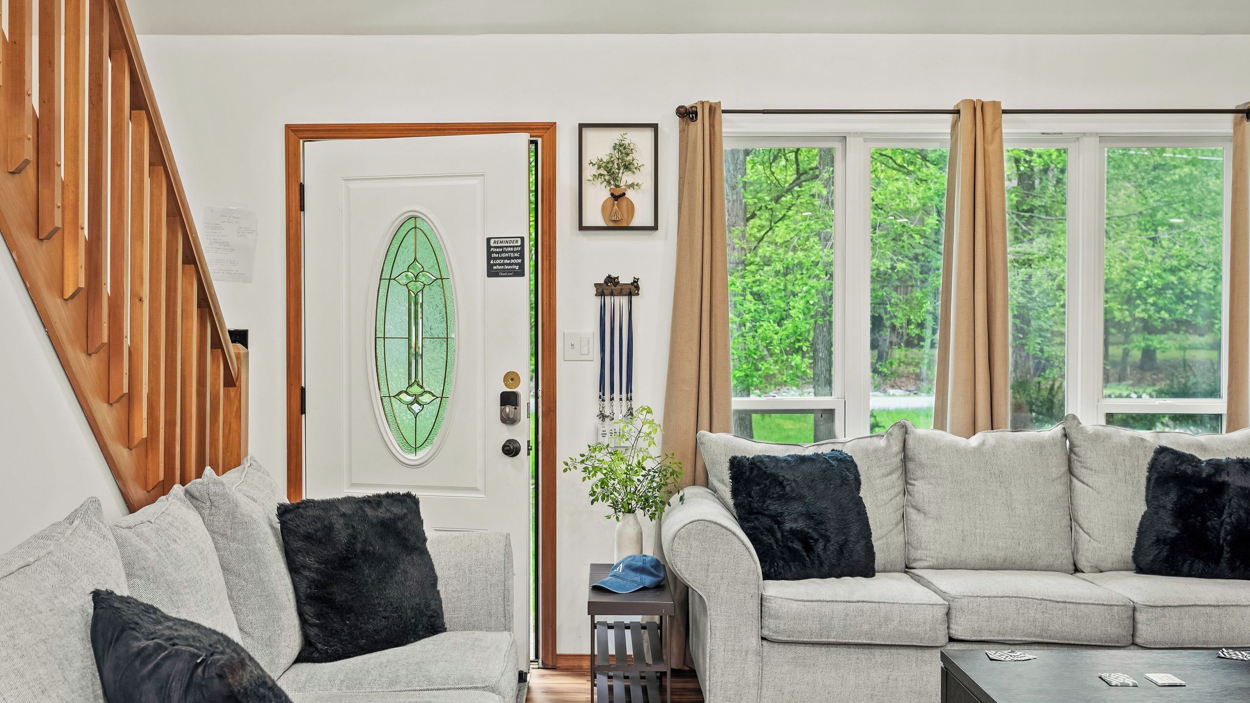 A living room with a staircase, a door with a green glass panel, a couch with black cushions, and a window with a view of greenery.