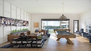 A spacious living room with a pool table, comfortable seating, a bookshelf, and a large window offering a view of the outdoors.