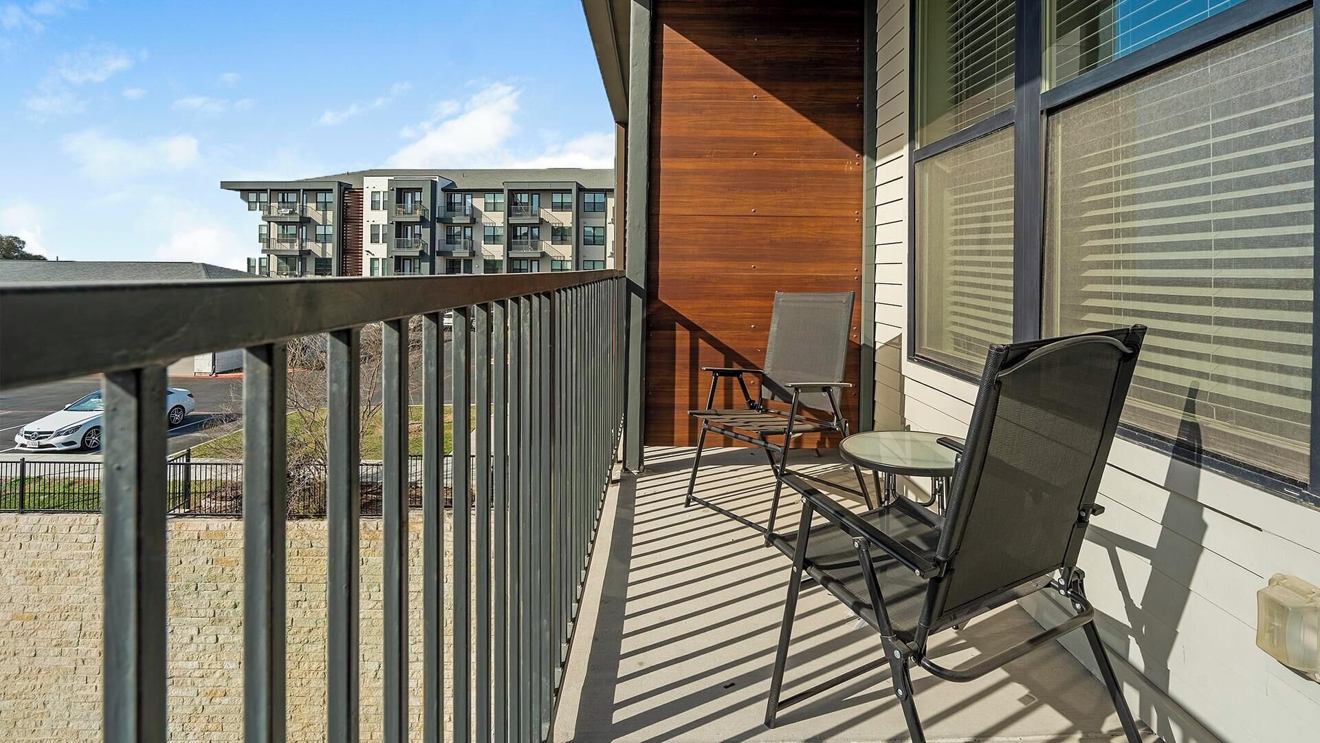 Ein Balkon mit Metallgeländer, einem Tisch und zwei Stühlen. Im Hintergrund ein mehrstöckiges Gebäude mit Balkonen.