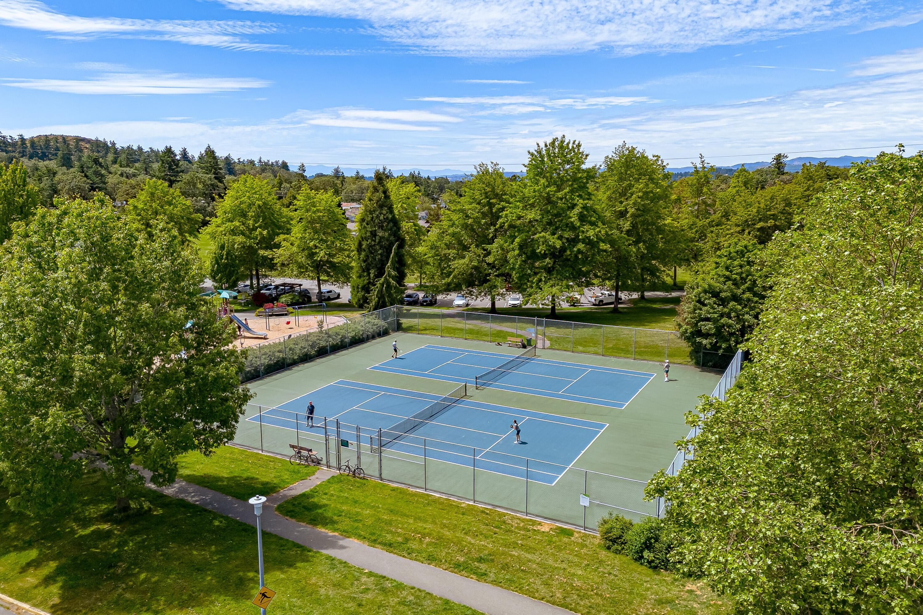 A tennis court surrounded by trees and a parking area.