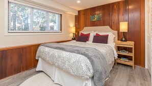 Een slaapkamer met een groot bed, houten lambrisering, een raam met jaloezieën en een nachtkastje met boeken.