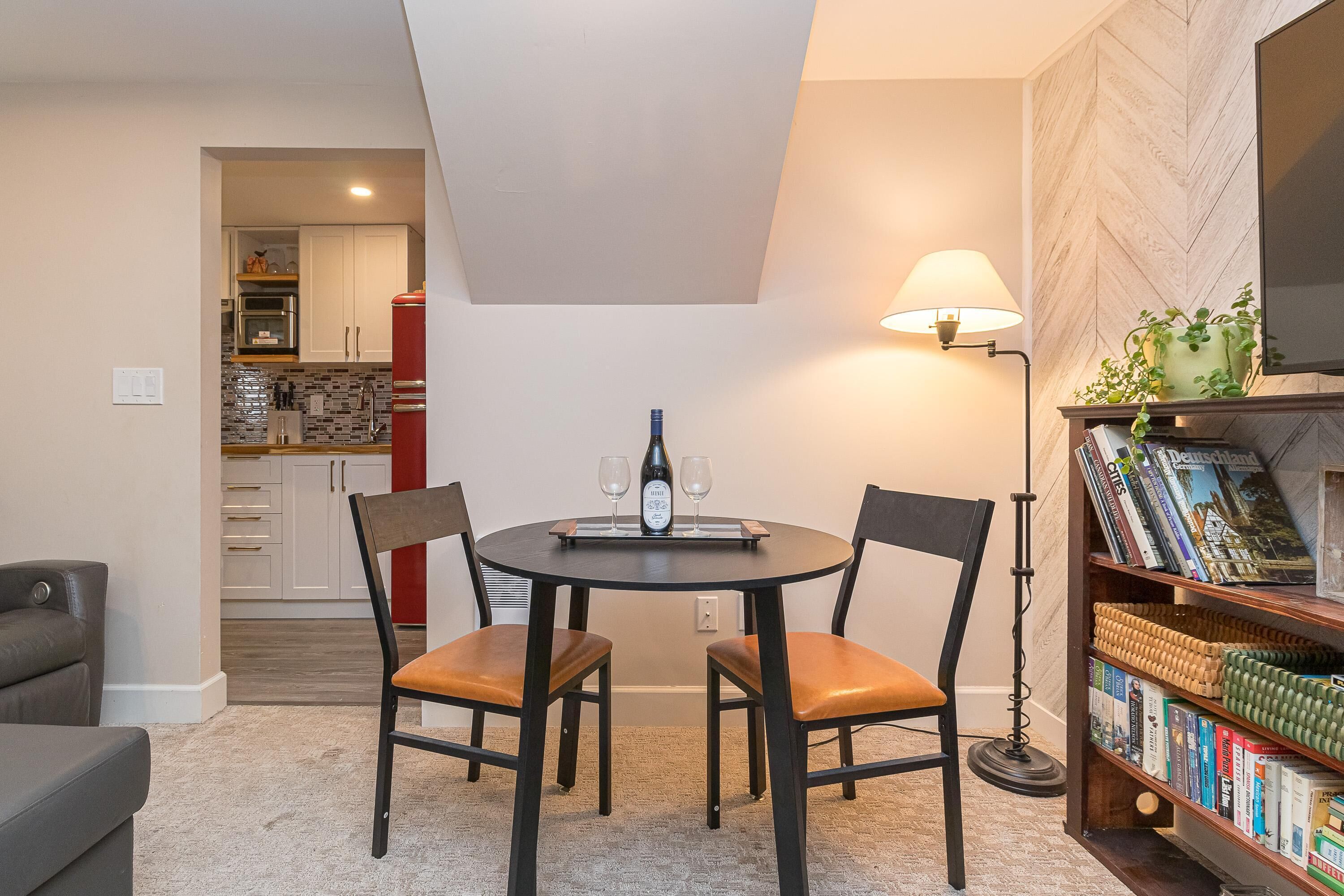 Un comedor con una mesa redonda, dos sillas, una botella de vino y dos copas. Se observa una estantería con libros y una lámpara. Se ve una cocina al fondo.