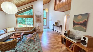 Un salon spacieux avec un plafond en bois, une grande fenêtre donnant sur les arbres, un canapé, une table basse, un tapis et une bibliothèque remplie de livres et d’un haut-parleur.