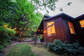A wooden cabin with a porch, surrounded by lush greenery and string lights. - Maple Fir Lodge - Hot Tub, Spacious, Fireplace, EV (Mount Hood Village)