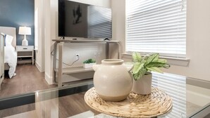 Ein modernes Wohnzimmer mit Flachbildfernseher, einem weißen Schrank, einer kleinen Pflanze und einer dekorativen Vase auf einem Glastisch.