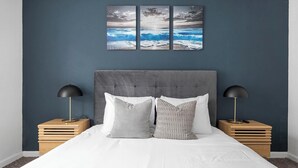 A bedroom with a grey upholstered headboard, white bedding, two bedside tables with lamps, and a wall-mounted triptych of ocean scenes. - Landing | Chic 1BD, Gym, Pool (Austin)