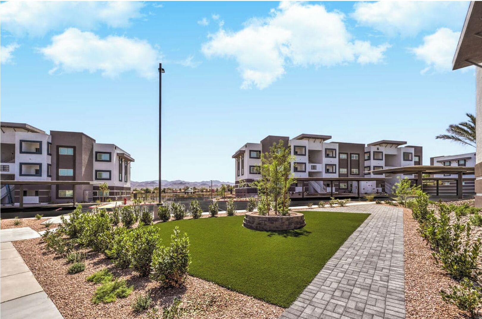 Un complexe résidentiel moderne comprenant plusieurs immeubles d’appartements, une pelouse centrale et un chemin piétonnier dallé.