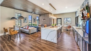 A modern kitchen with a central island, wooden flooring, pendant lighting, and a view of the outdoors. - Landing | Amazing 2BD, Rooftop Lounge, Pool (Henderson)