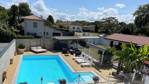 Una piscina rectangular con un borde empinado y azulejos, rodeada por una valla blanca. Hay sillones de descanso y una pequeña mesa con toldo.