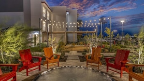 An outdoor seating area with red chairs and a circular table, surrounded by greenery and a building with string lights.