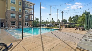 A swimming pool with lounge chairs and umbrellas, surrounded by a building with multiple floors and a brick facade.