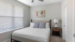Une chambre à coucher avec un lit, une table de chevet, une lampe, une fenêtre avec des stores et un tableau au mur.