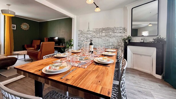 Un comedor con una mesa de madera dispuesta para una comida, un televisor al fondo y un mural con un paisaje urbano en la pared.