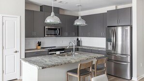 A modern kitchen with dark grey cabinets, stainless steel appliances, a marble countertop, and two pendant lights. - Landing | Amazing 2BD, Gym, Clubhouse (Huntsville)