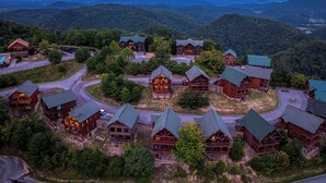 A cluster of log cabins with a winding road connecting them, surrounded by forested hills. - OMG VIEWS! Gameroom~Hot tub~Outdoor Pool~ Suana (Sevierville)