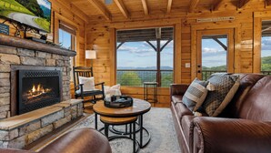 A cabin interior with a stone fireplace, wooden walls, a leather couch, and a large window offering a scenic view. - OMG VIEWS! Gameroom~Hot tub~Outdoor Pool~ Suana (Sevierville)