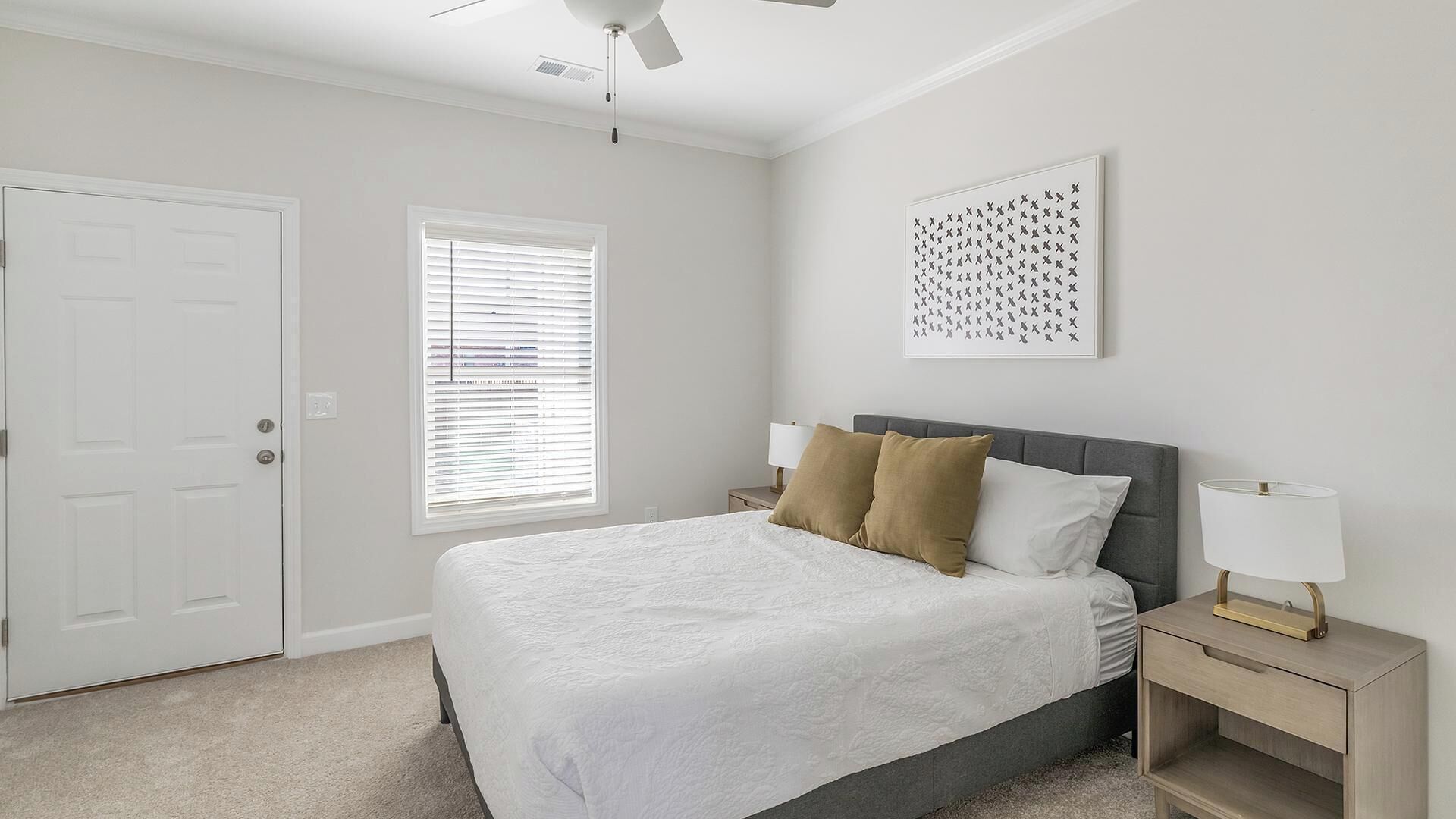 A bedroom with a bed, a nightstand, a wall-mounted artwork, a ceiling fan, and a door.
