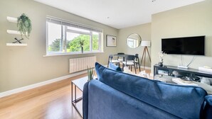 A modern living room with a blue sofa, a dining table with chairs, a TV, and a large window with a view of greenery. - Beautiful apartment in Claygate Village (Claygate)