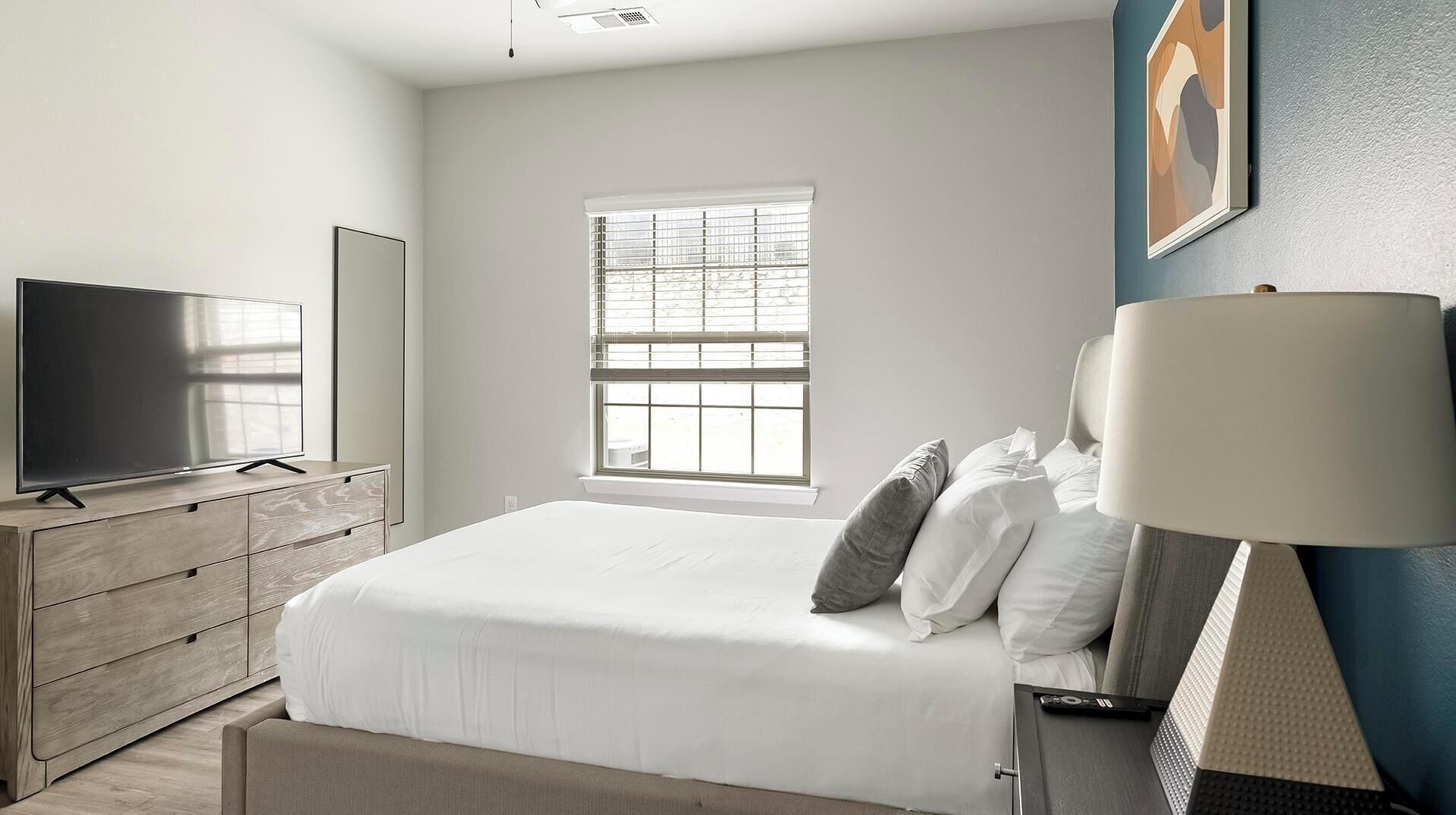 A bedroom with a bed, a TV on a dresser, a window, and a lamp.