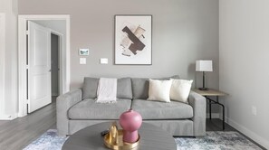A living room with a grey sofa, a round coffee table with a pink vase, a lamp, and a framed abstract artwork on the wall.
