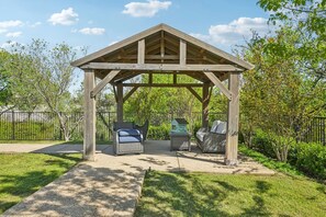 A wooden gazebo with two wicker chairs and a small table, surrounded by a fenced garden.