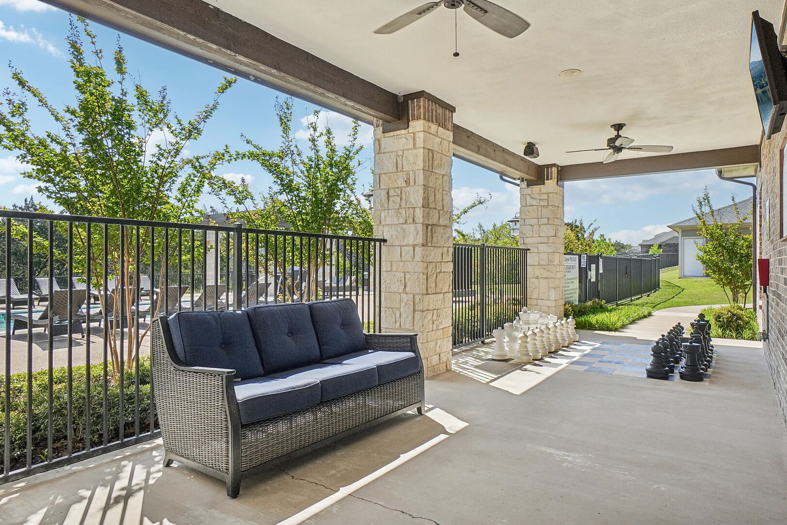 A covered patio with a wicker sofa, stone pillars, a ceiling fan, and a view of a backyard with a pool and greenery.