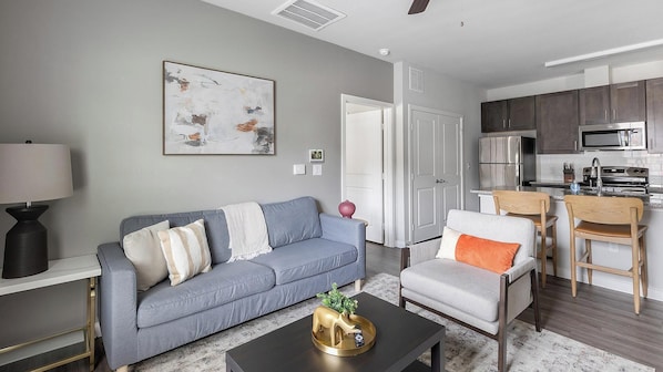 A modern living room with a grey sofa, a coffee table with a decorative animal, and a kitchen area with dark cabinets and stainless steel appliances. - Landing | Stylish 1BD, Gym, Pool (Dallas)