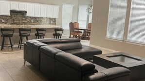 Una sala de estar moderna con un sofá seccional, un comedor con mesa y sillas, y una cocina con armarios blancos y un salpicadero de piedra.