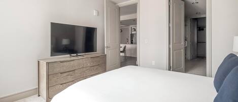 Ein Schlafzimmer mit einem großen Bett, einem Fernseher auf einer Holzkommode und Blickrichtung zu einem Badezimmer und einer Küchenzeile.