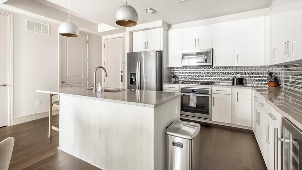 Una cocina moderna con armarios blancos, electrodomésticos de acero inoxidable, una isla central con pileta e iluminación colgante.