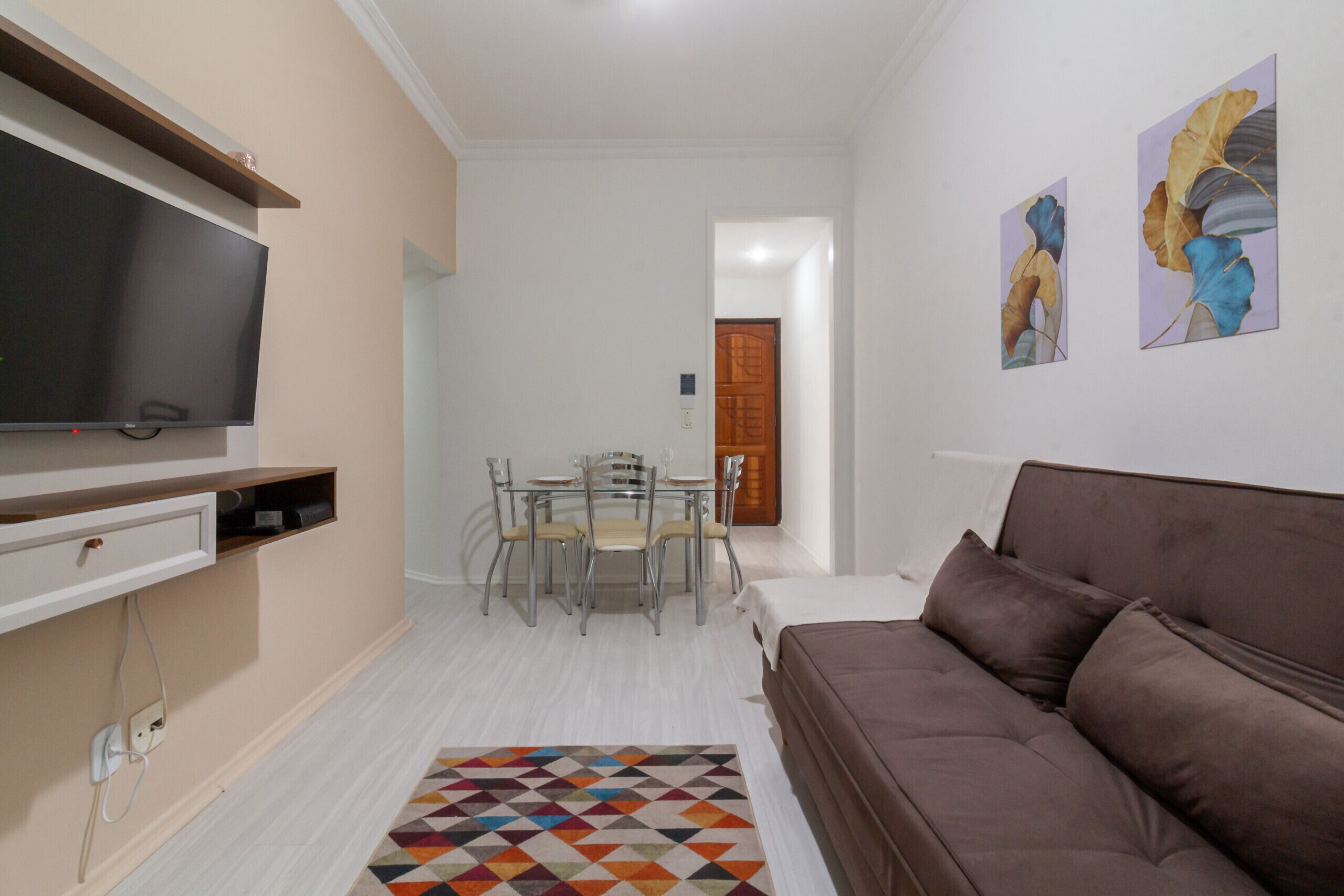 A modern living room with a flat-screen TV, a brown sofa, a dining table with chairs, and two wall-mounted artworks.