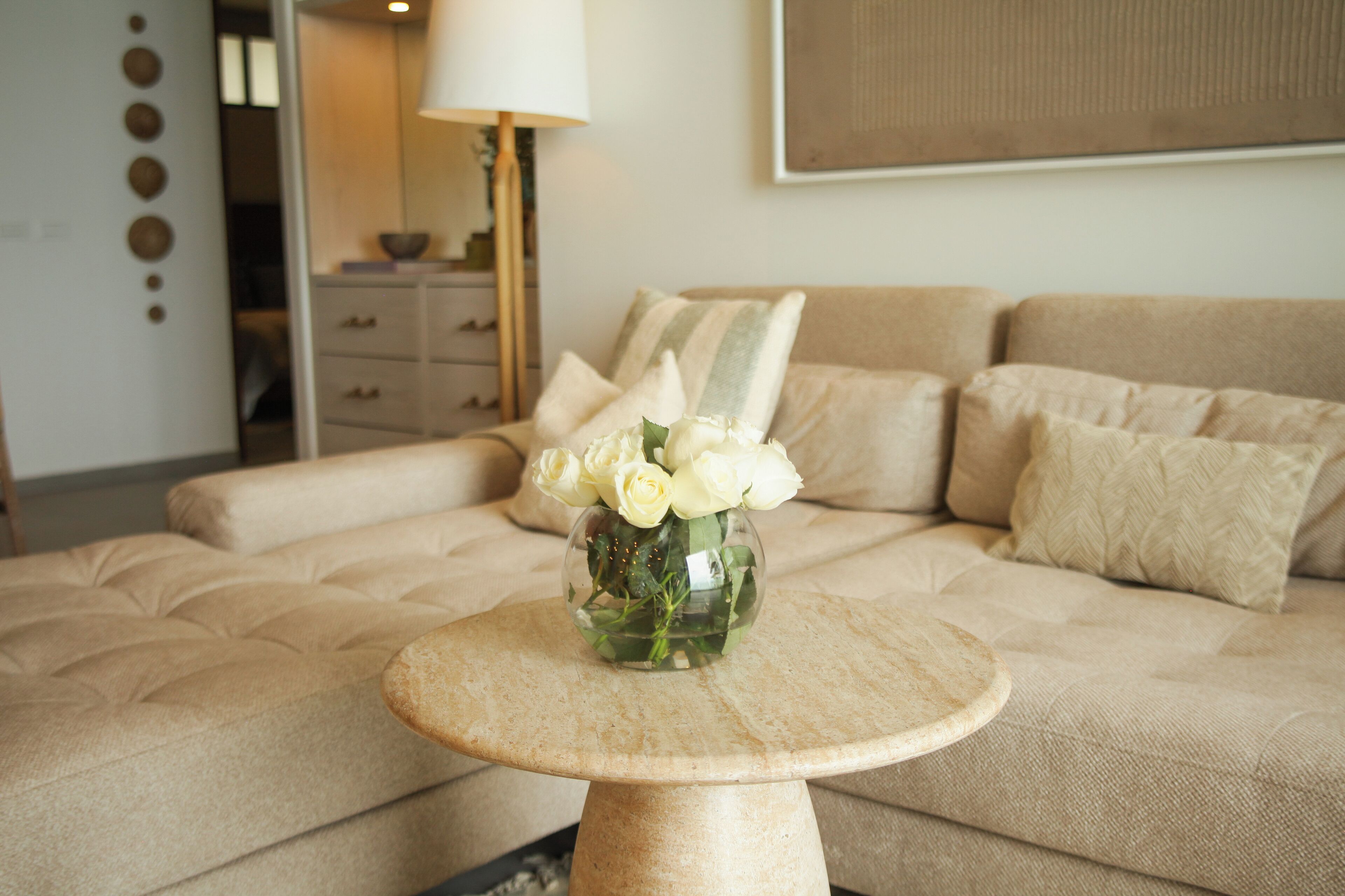 Una sala de estar con un sofá beige, una mesa de centro redonda y un jarrón con rosas blancas.