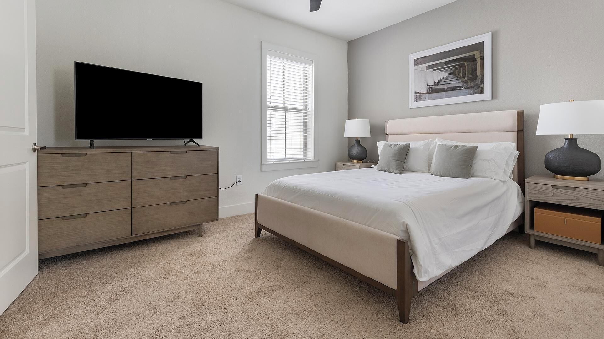 A bedroom with a bed, a dresser, a TV, a window, and a nightstand with a lamp.